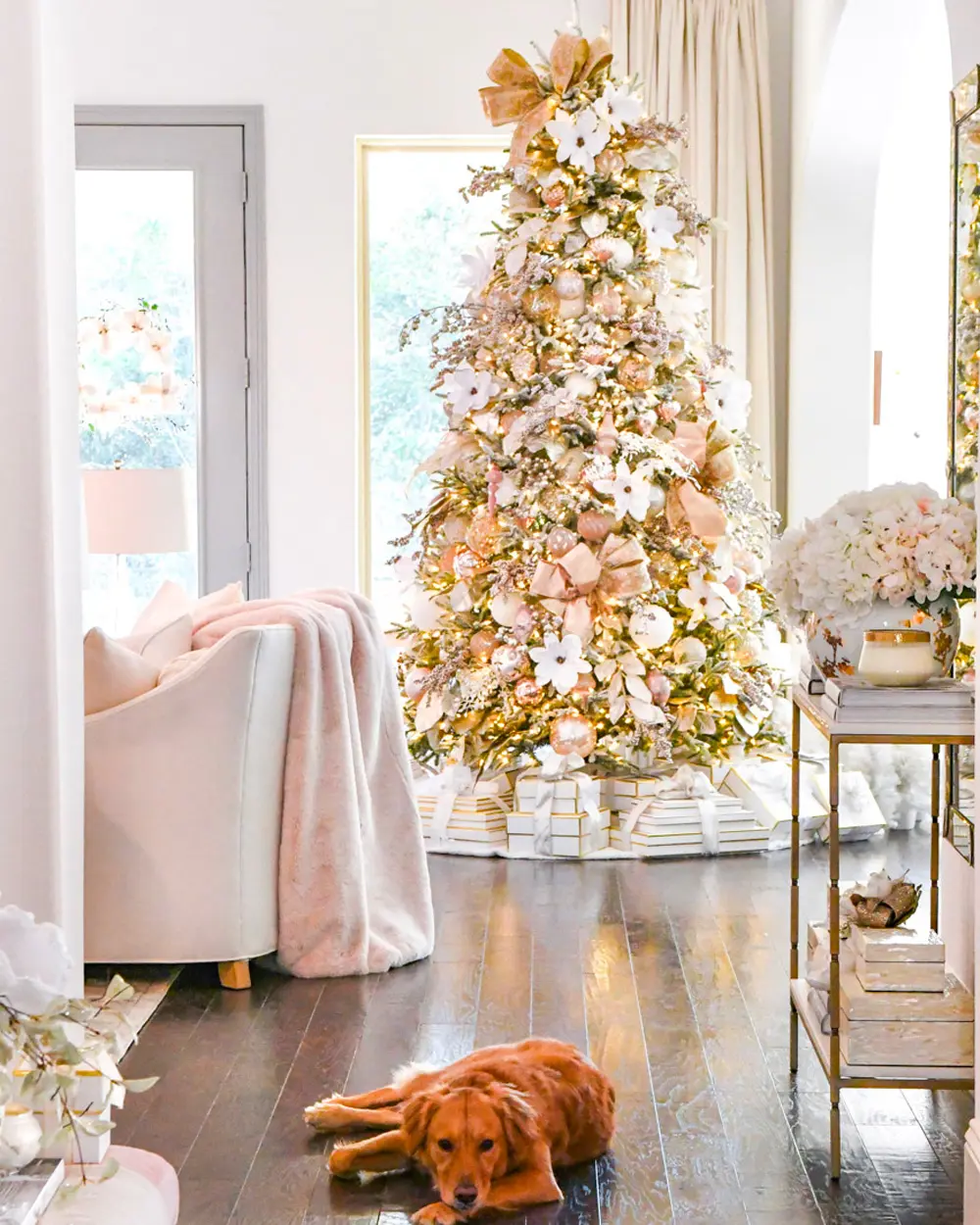Flocked tree decorated in blush pink and gold with ribbons, near a pink chair and a sleeping dog
