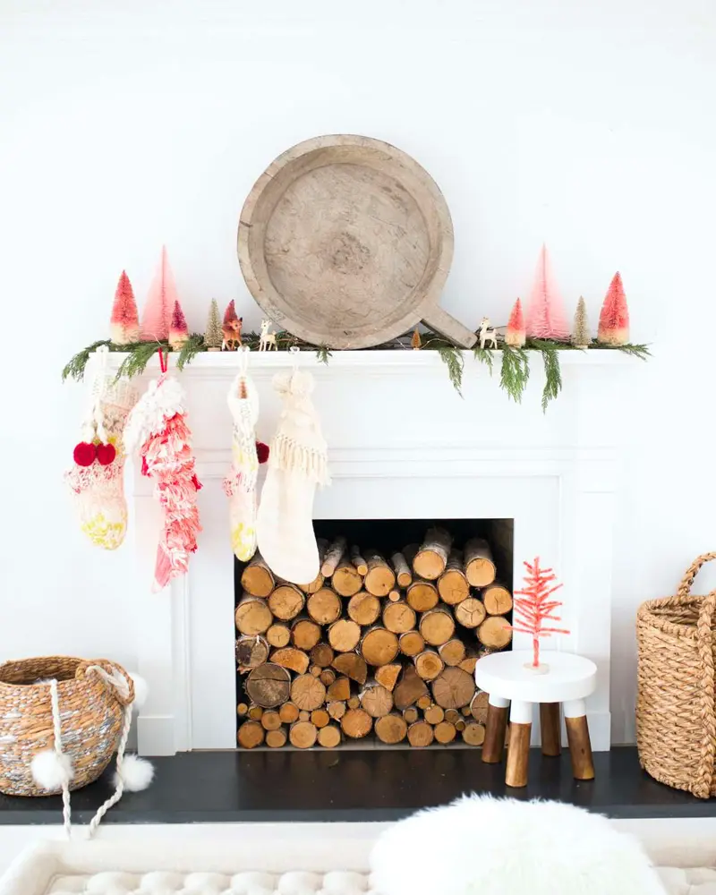 White fireplace with stacked logs, pink and white knit stockings, small pink bottle-brush trees and a round wooden tray on the mantel