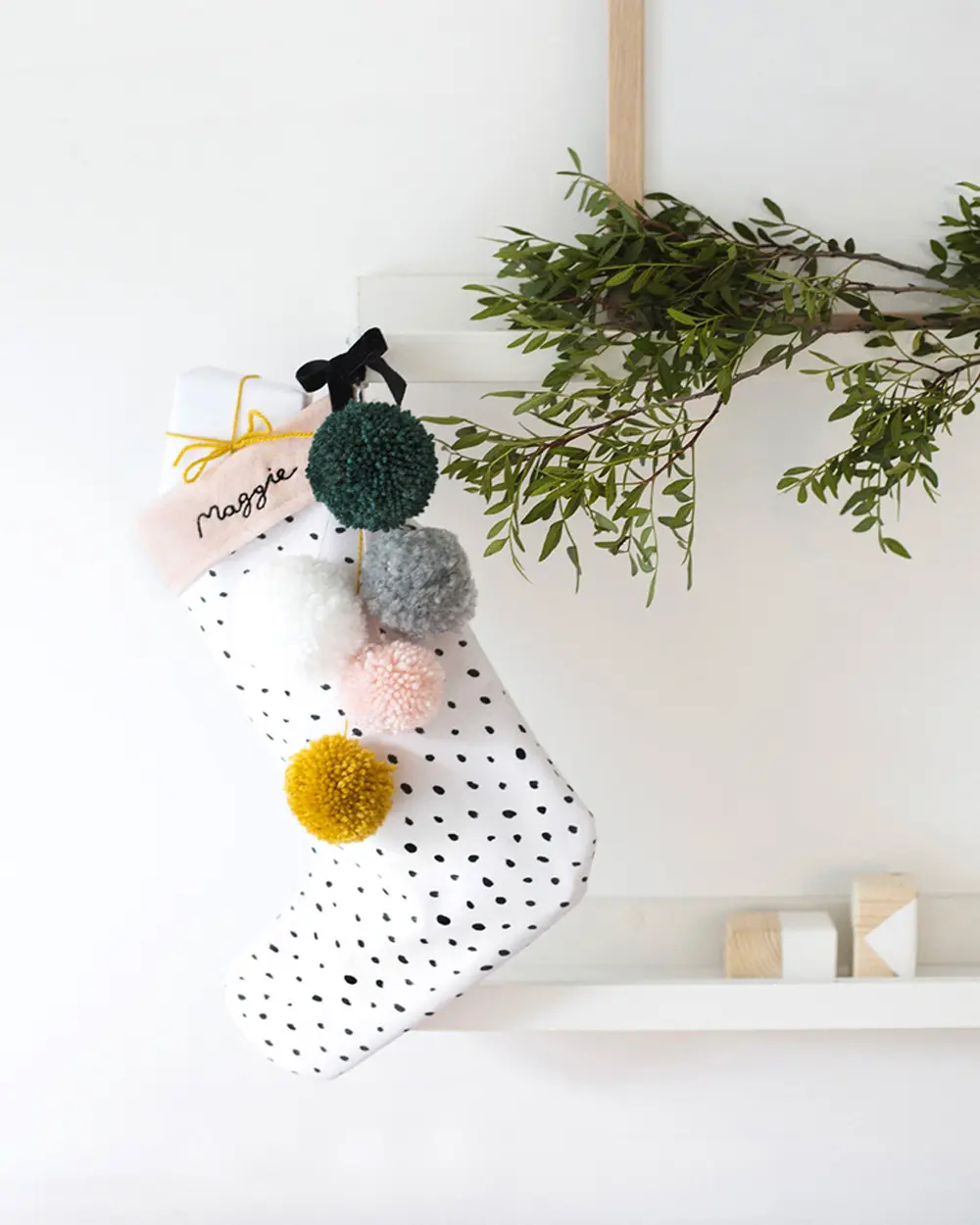 White stocking with black dots, colorful yarn pompoms and a name tag hanging from a branch