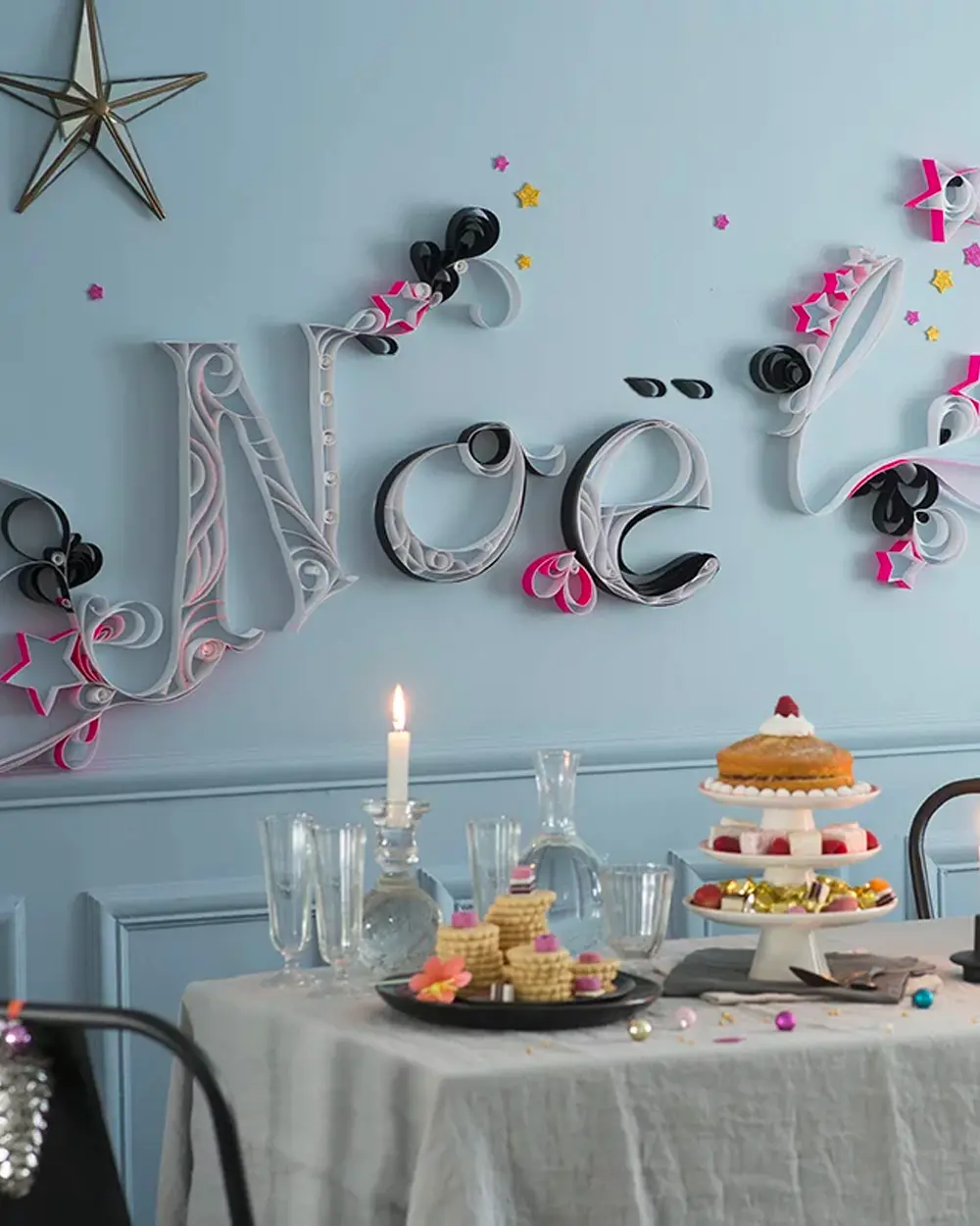 Quilled paper letters spelling Noël with paper stars on a blue wall above a dessert table