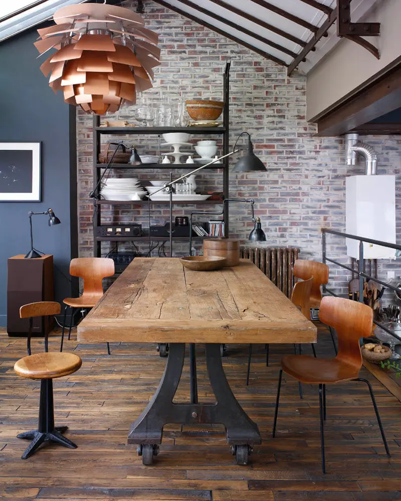 Reclaimed plank table on a cast-iron base with orange chairs, a copper pendant and brick wall