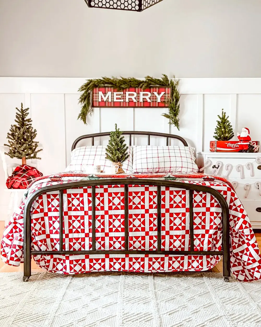 Iron bed with a red and white quilt, a Merry sign with greenery and small trees on nightstands