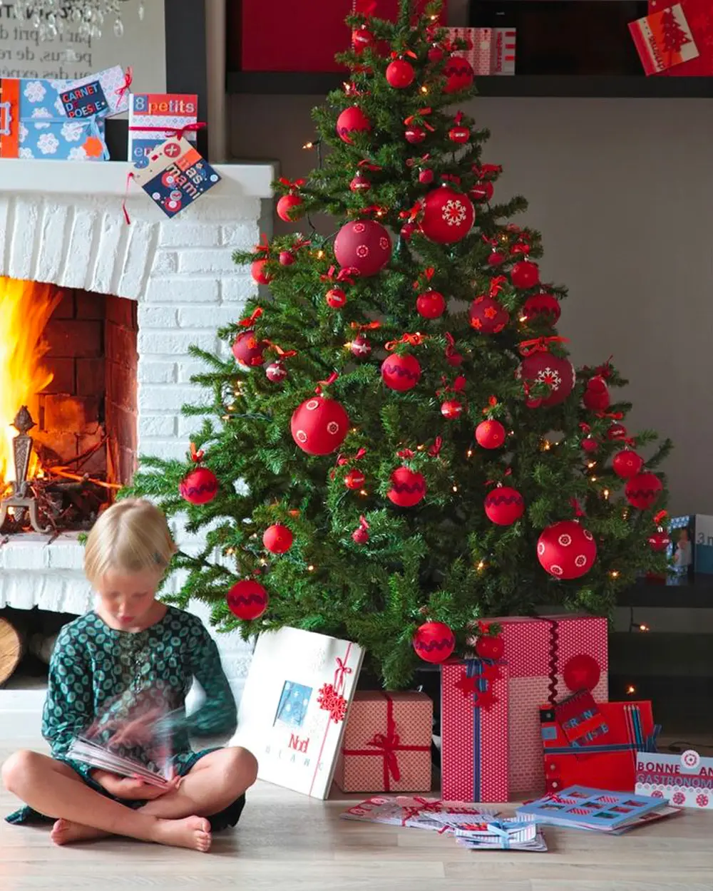 Child reading under a green tree covered in red ornaments, with red presents and a fireplace