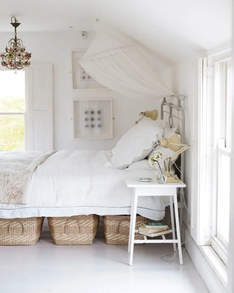 All-white bedroom with a draped canopy, a beaded chandelier and baskets under the bed