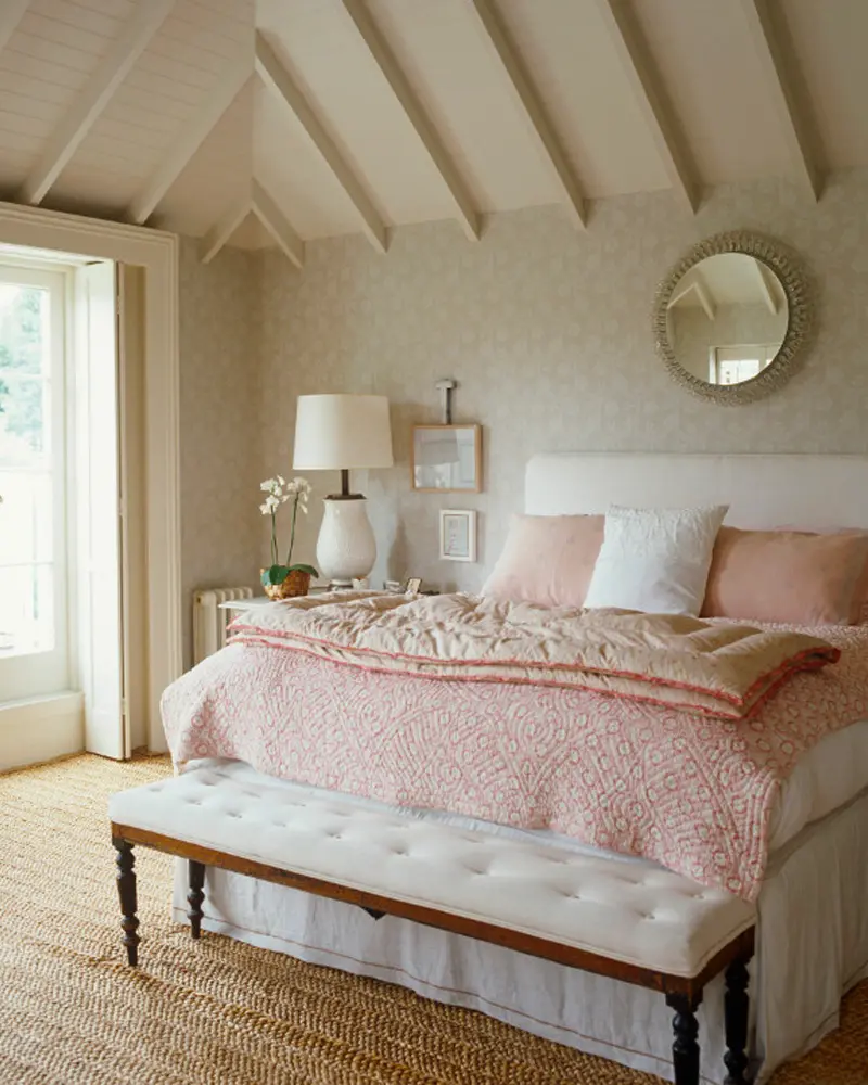 Pink quilted bedspread, a tufted bench, a round mirror and a vaulted ceiling