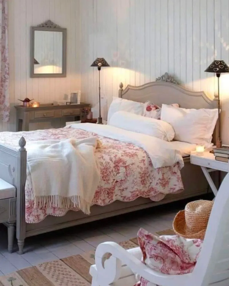 Pink floral bedding on a gray painted bed, a white rocking chair and twin lamps on paneled walls