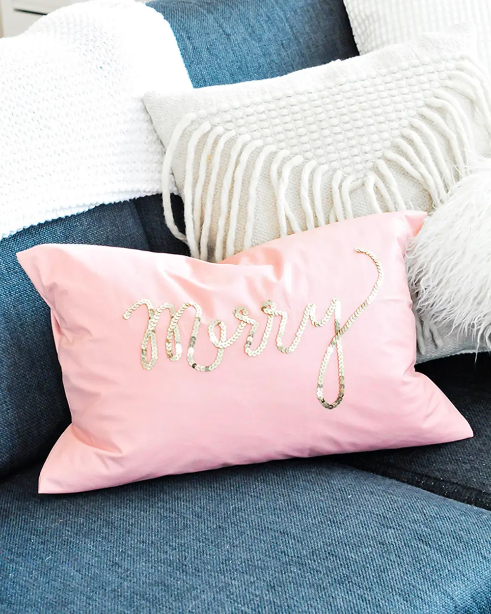 Pink velvet lumbar pillow with gold sequin script reading Merry on a blue sofa