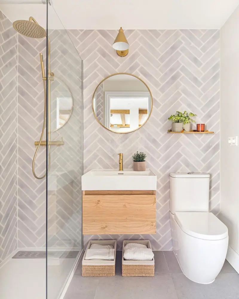Pale herringbone tile wall with round brass mirror, small wood vanity and brass shower fixtures