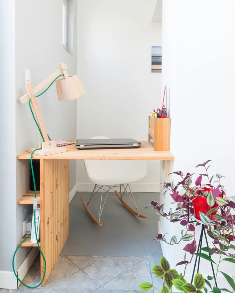 Small wood desk with pink lamp and rocking chair by a white window wall