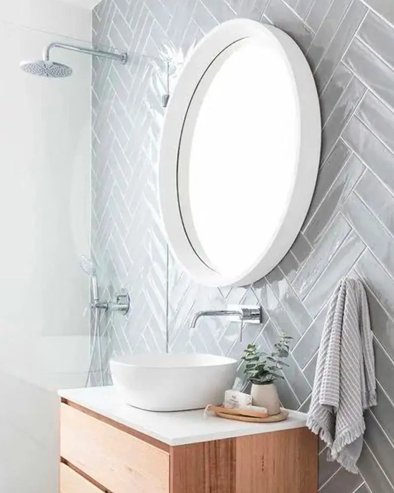 Gray herringbone tile wall, round white mirror, wood vanity with white basin and gray towel