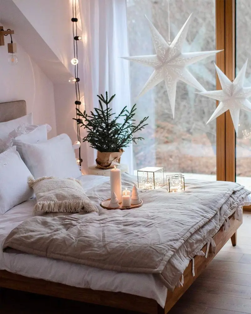 Low bed with linen bedding, small potted fir, candles and paper star lamps in the window