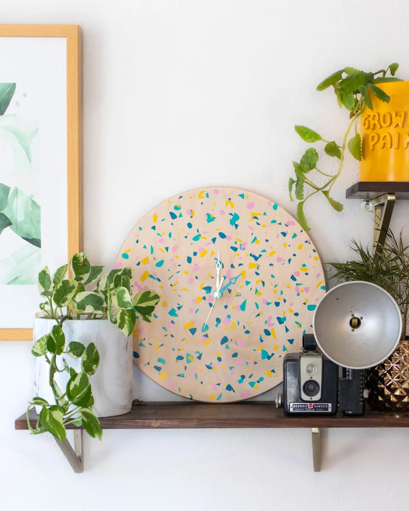 Round wall clock with colorful vinyl chips on a shelf with plants and a vintage camera