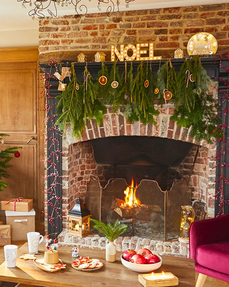 Brick fireplace with a fir garland, dried orange slices, NOEL marquee letters and a lit fire