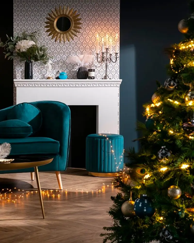 Living room with teal velvet armchair and pouf, sunburst mirror and lit tree