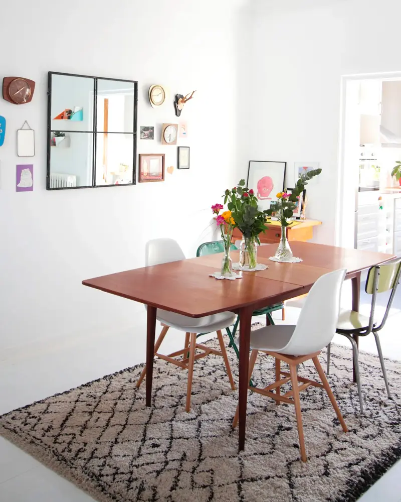 Teak dining table with white molded chairs on a Berber rug, flowers and a gallery of small frames
