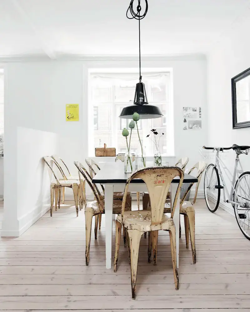 White table with worn gold metal chairs, a black pendant and a bicycle against white walls