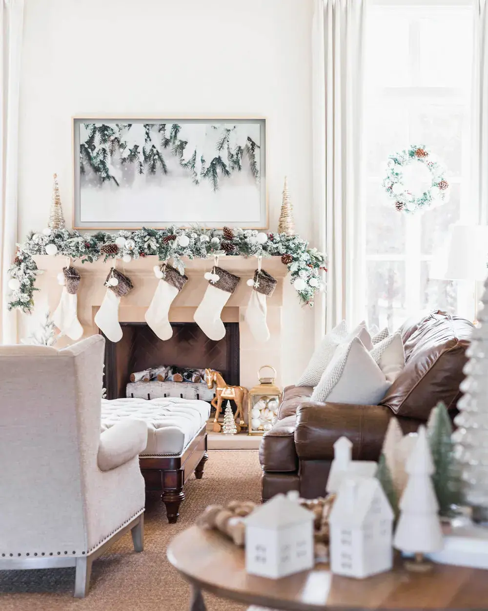 Chic living room with a frosted garland and white stockings on the mantel and little white ceramic houses on the coffee table