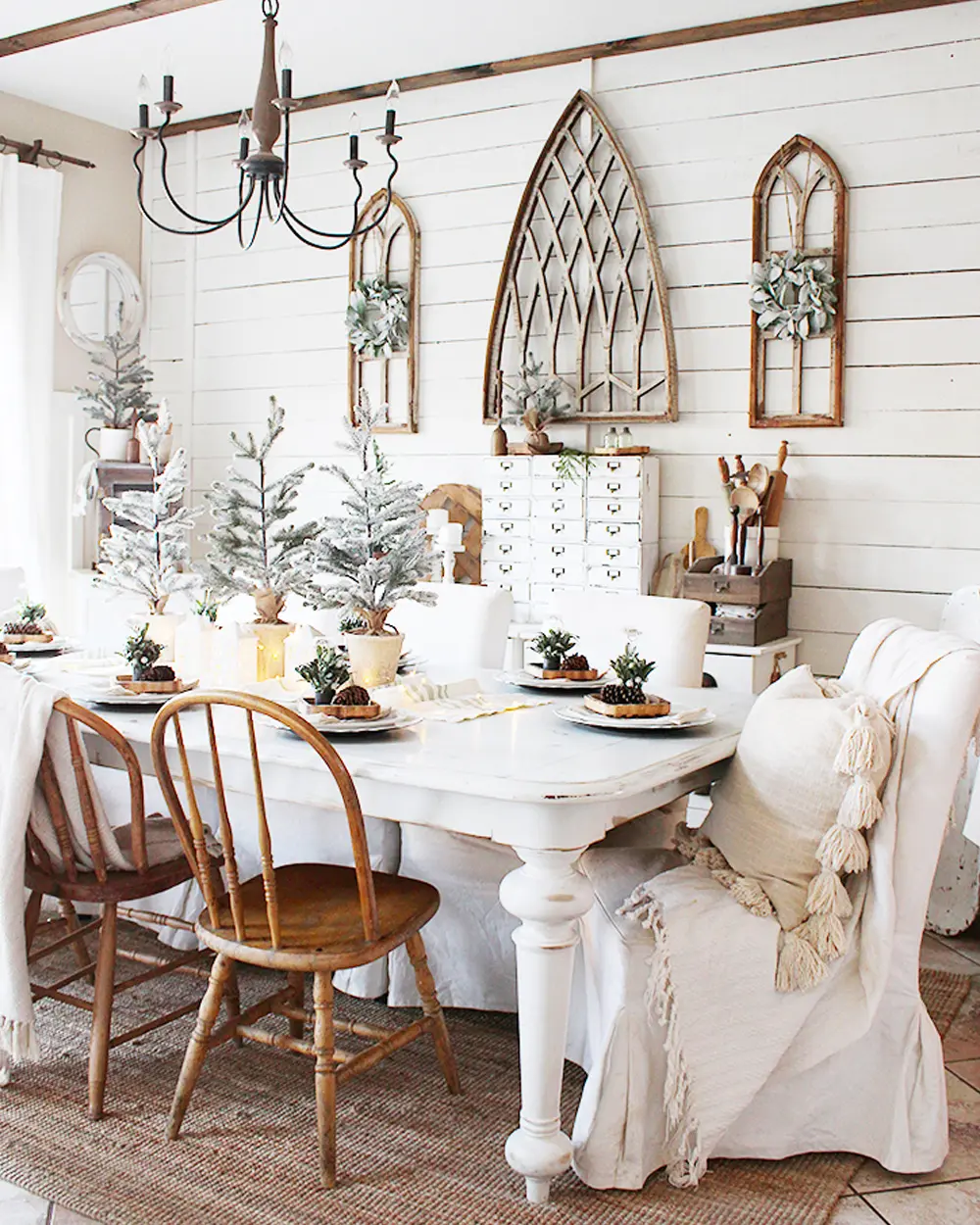 White farmhouse table with mini flocked trees, mismatched wood chairs, shiplap wall and arched window frames