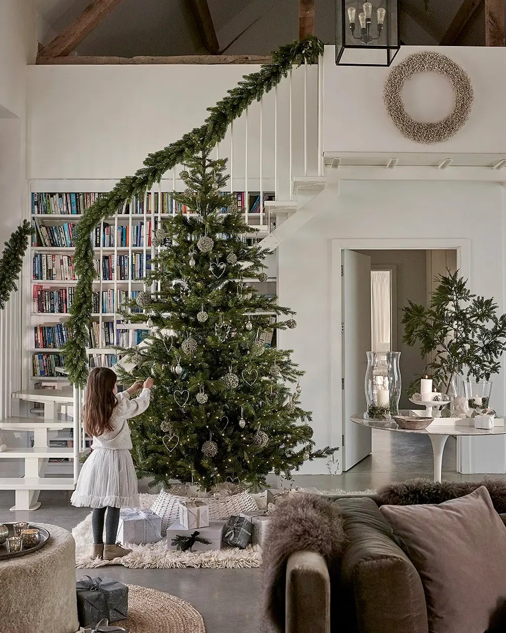 Child decorating a tall green tree with silver ornaments below a staircase wrapped in a fir garland