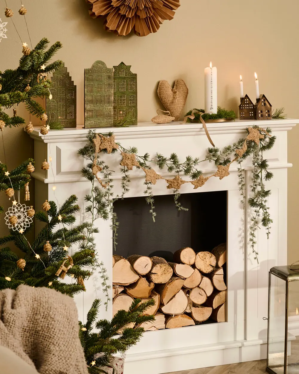 White mantel with a trailing greenery garland, star garland, candles, green paper houses and stacked firewood