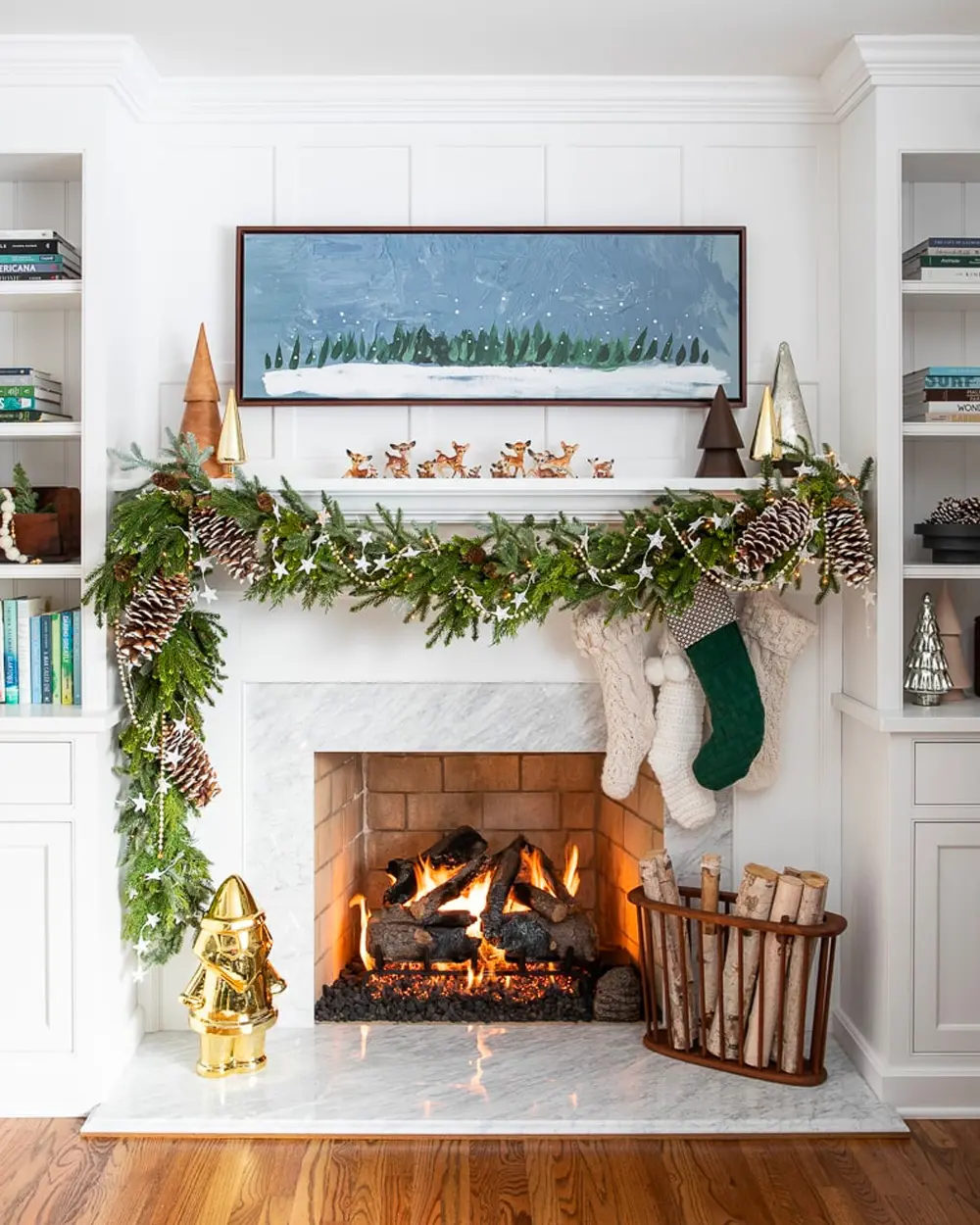 White fireplace with a lush greenery and pinecone garland, knit stockings, built-in shelves and a winter landscape painting