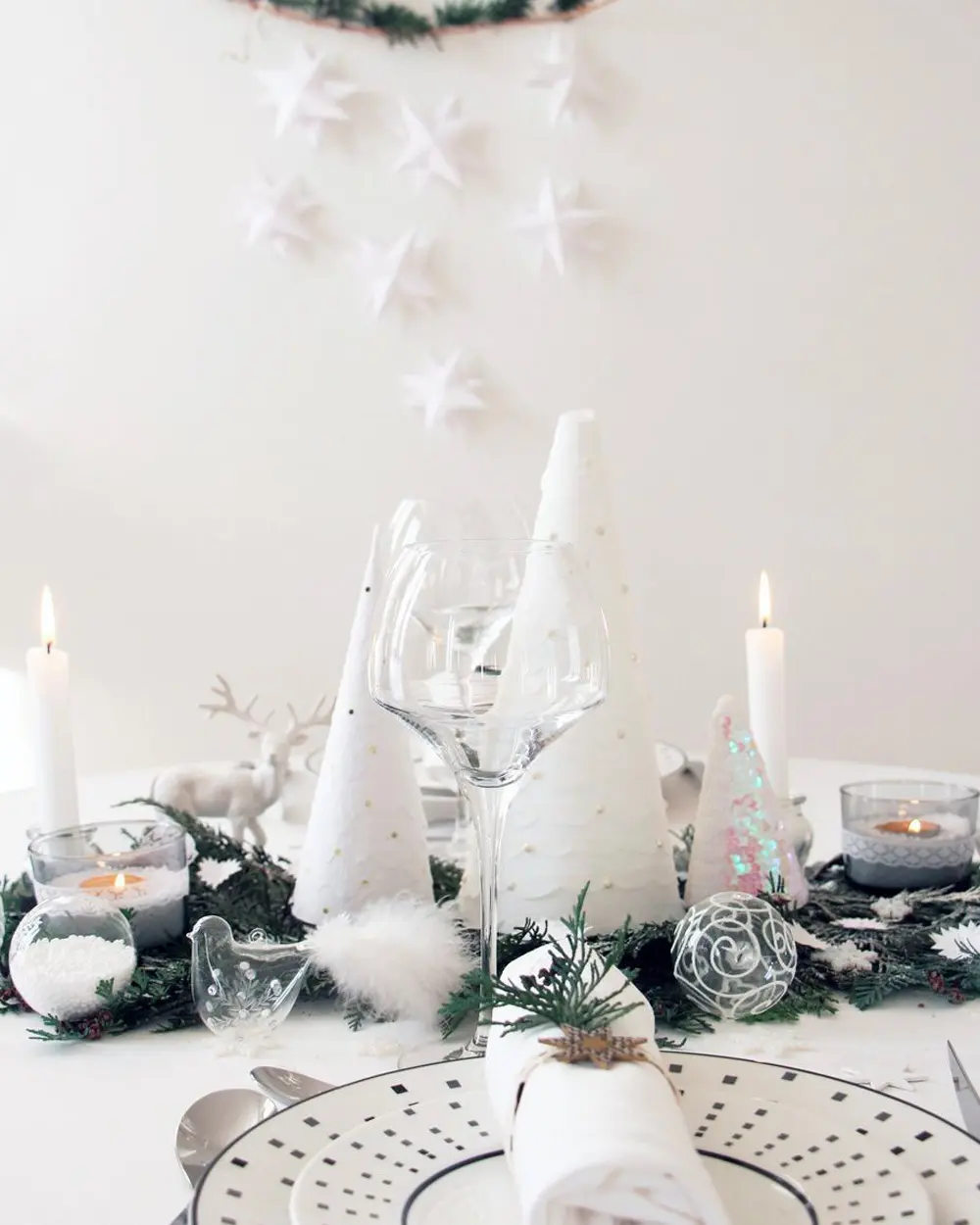 Christmas table with white cone trees, candles, glassware and a fir runner, with paper stars hanging above