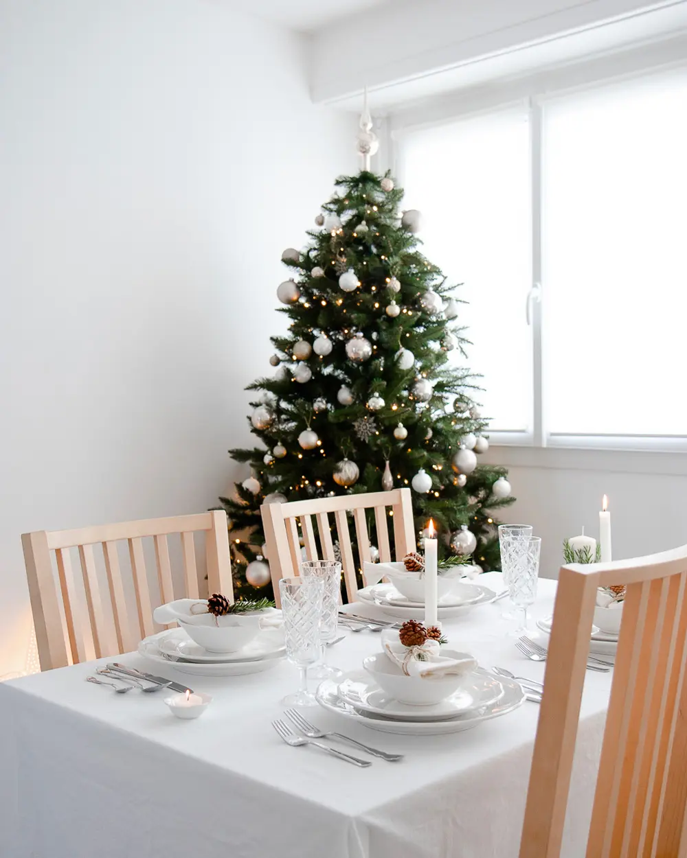 Minimalist dining room with a white tablecloth, white plates, light wood chairs and a tree with silver ornaments