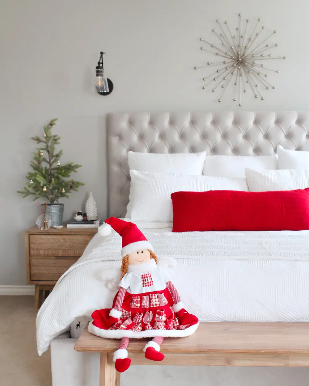 Tufted white headboard, white bedding with a red pillow, a small potted tree and a festive doll on a wooden bench