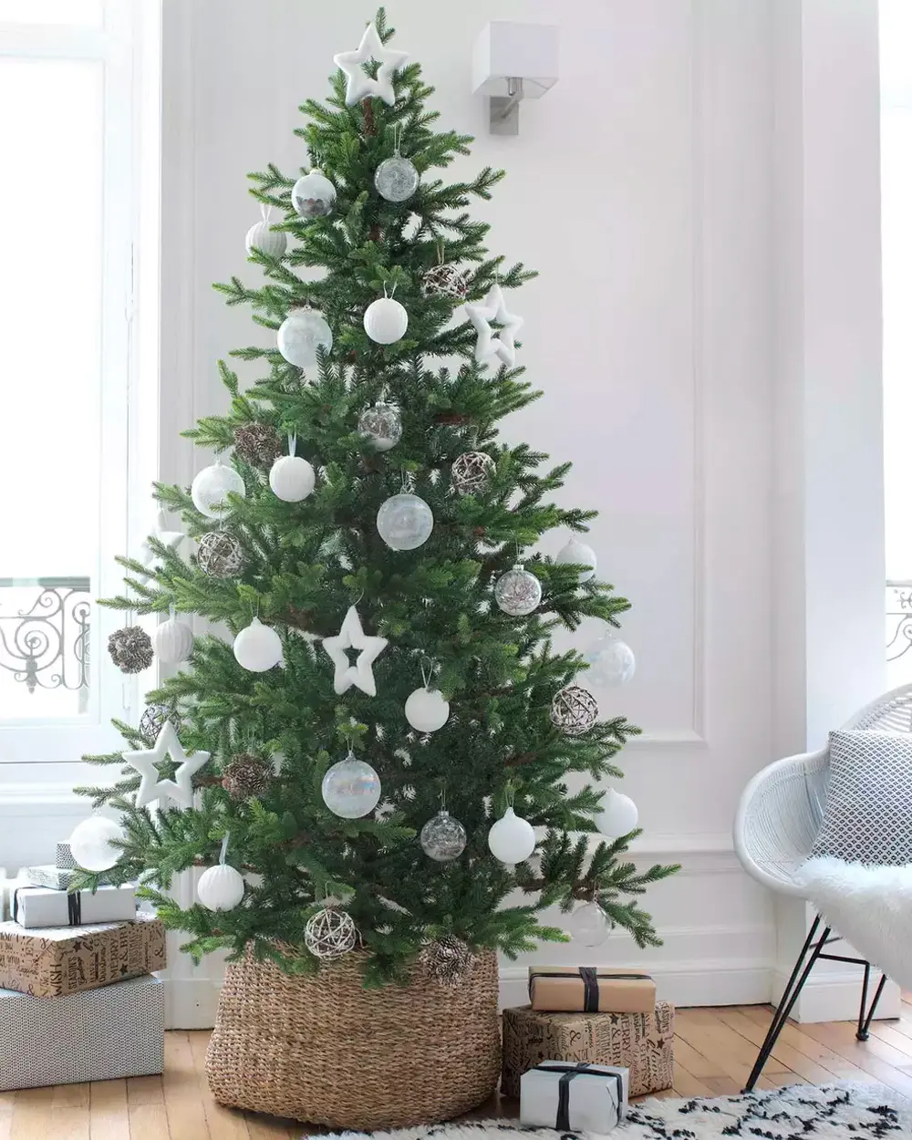 Green Christmas tree trimmed with white baubles and stars, set in a woven basket by a tall window