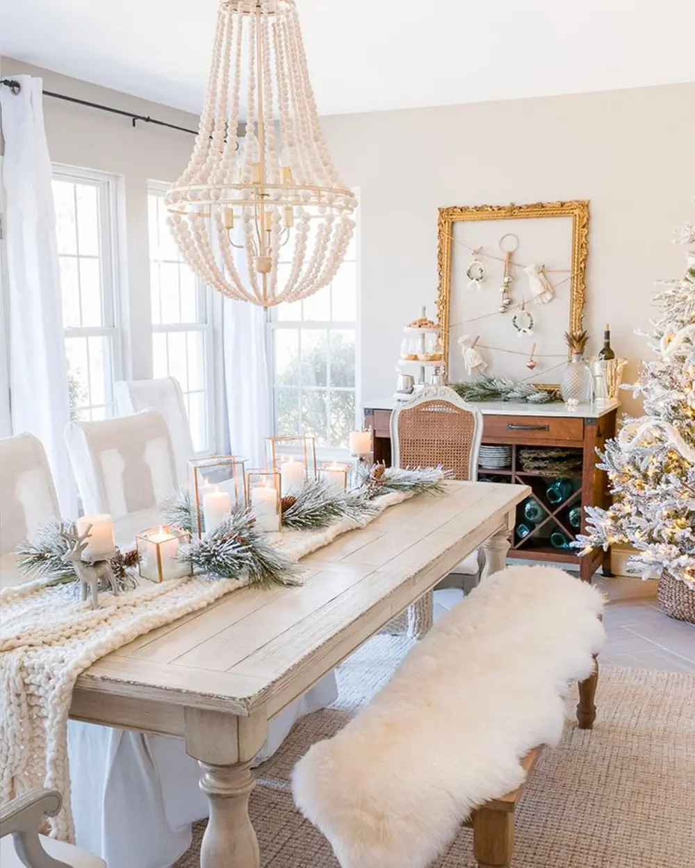 Long pale wood table with a greenery runner and candles, a beaded chandelier, sheepskin-draped bench and flocked tree