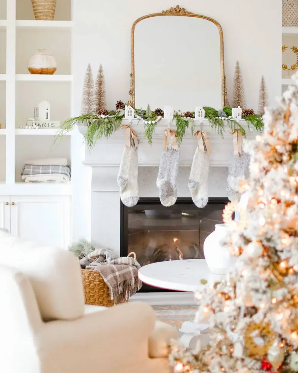 White fireplace with a gold arched mirror, greenery garland, gray knit stockings and a flocked tree with warm lights