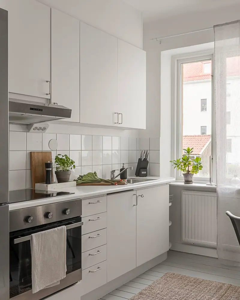 Narrow white kitchen with square tile backsplash, wood cutting board and potted plants by the window