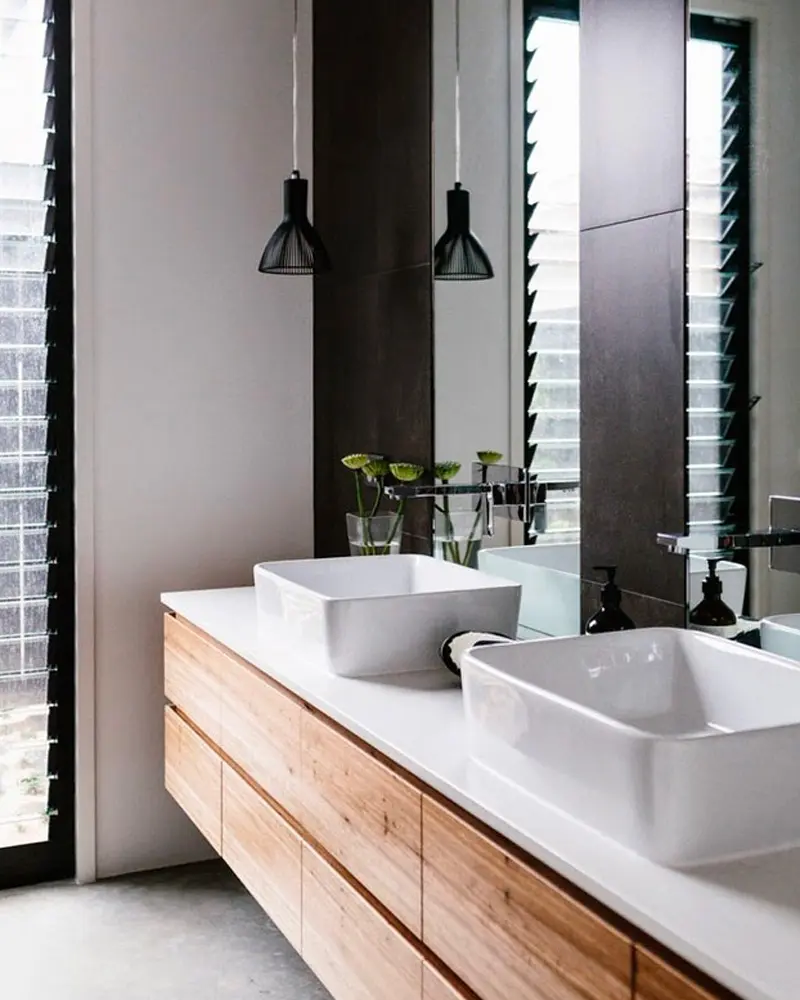 Floating wood double vanity with white vessel sinks, black pendants and dark louvered window