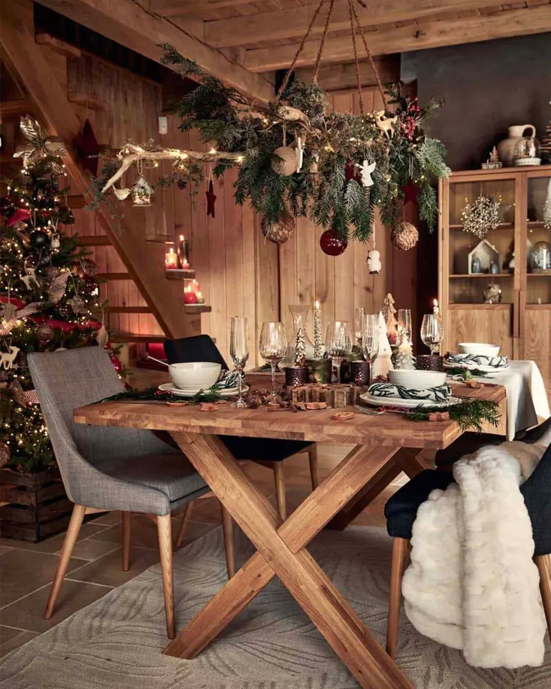 Trestle wood dining table under a hanging greenery chandelier with ornaments in a wood-lined room
