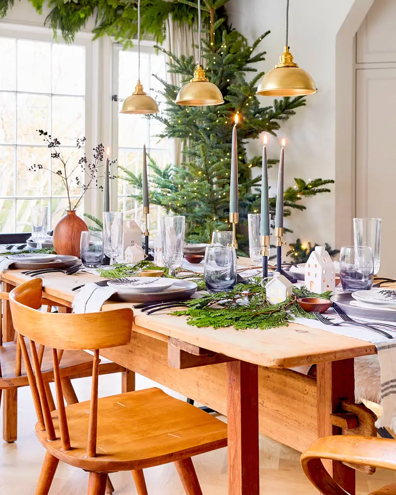 Wooden dining table with a fir runner, taper candles and brass pendants in front of a big indoor tree