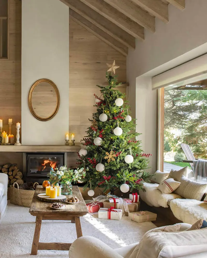 Tree with white baubles and red berries in a living room with pale wood walls, a beamed ceiling and a lit fireplace