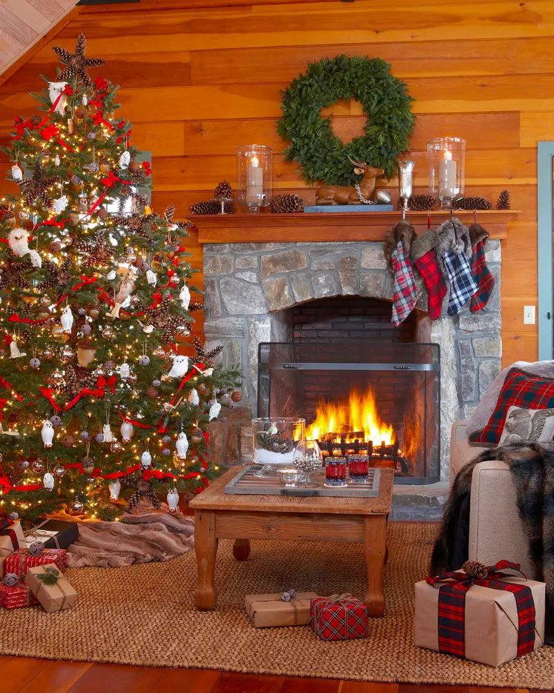 Stone fireplace in a wood-paneled room with a wreath, plaid stockings, a tree with red ornaments and a lit fire