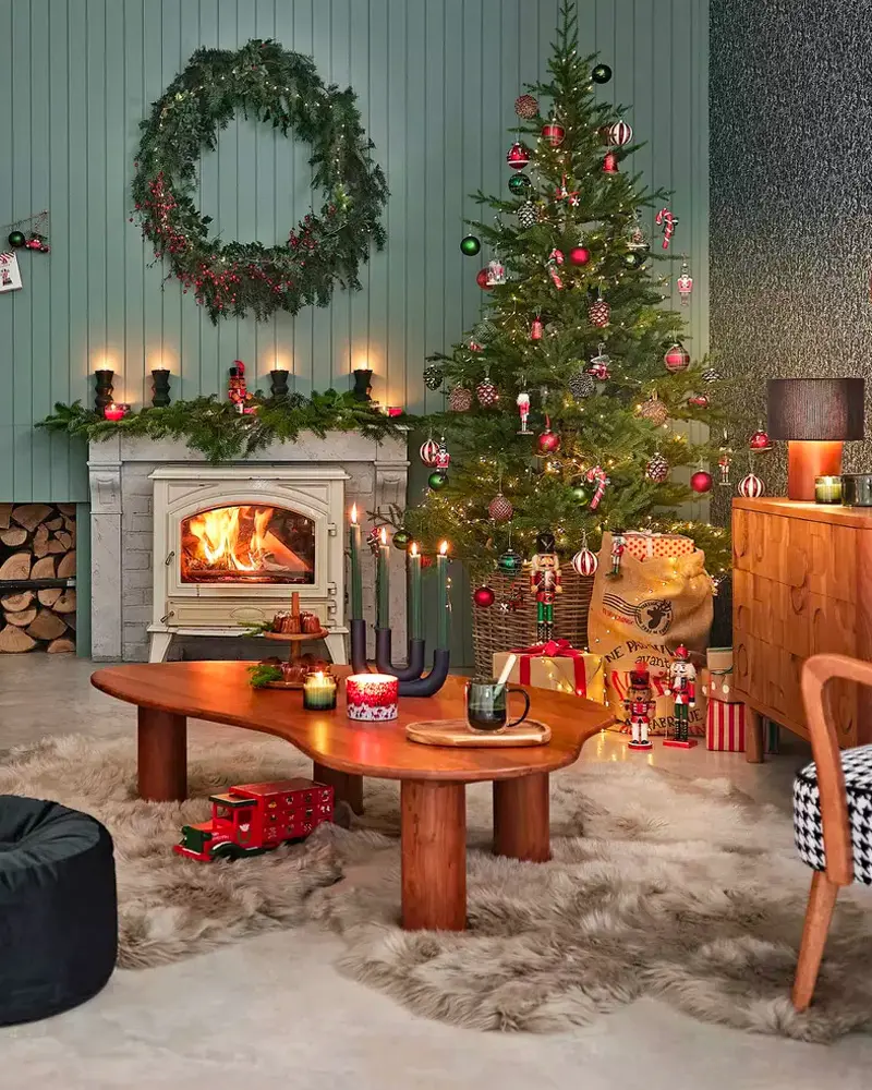 Wood stove, a curvy wooden coffee table, a red toy truck and a tree with red ornaments against a sage wall