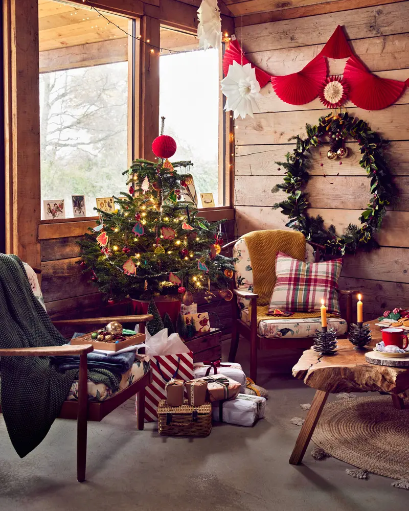 Wood-paneled cabin room with a small lit tree, plaid pillows, a wreath and a red felt banner