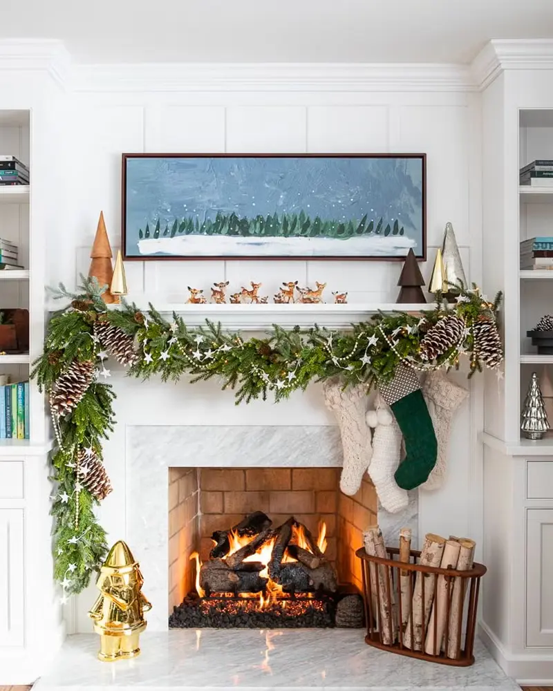 White fireplace with a pine cone garland, gold trees, stockings and a snowy landscape painting