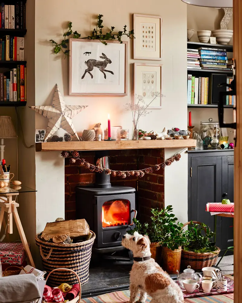 Wood stove under a beam mantel with a felt ball garland, a wooden star, framed prints and a terrier