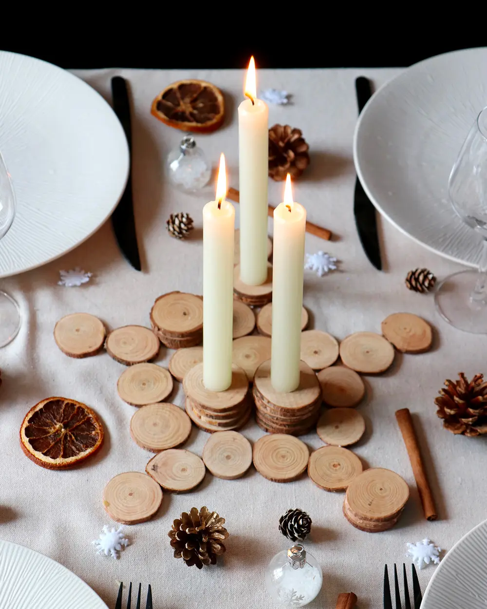 Centerpiece of three white taper candles on a scatter of wood slices with dried oranges and pinecones