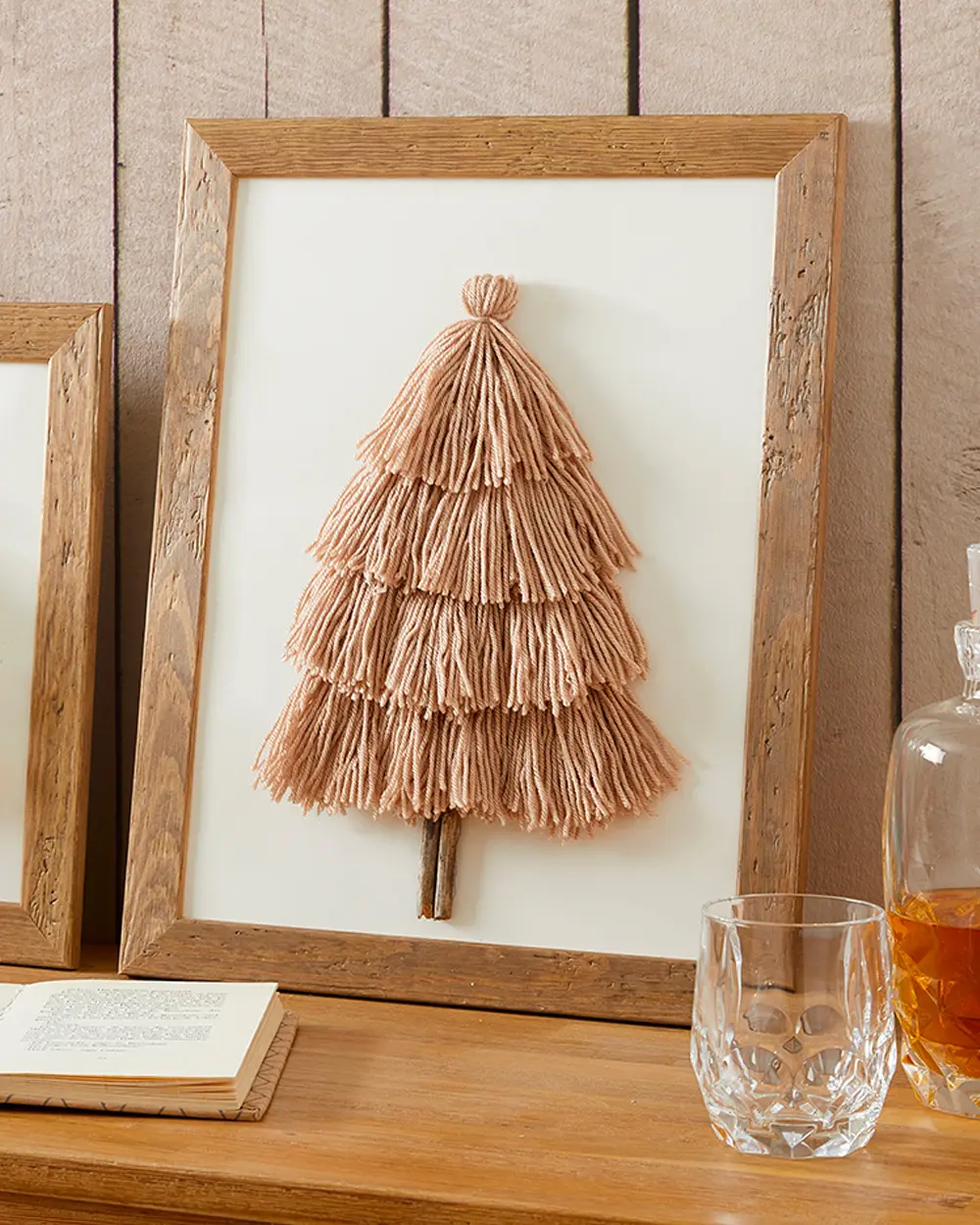 Framed wall art with a Christmas tree made of rows of beige yarn tassels and a twig trunk