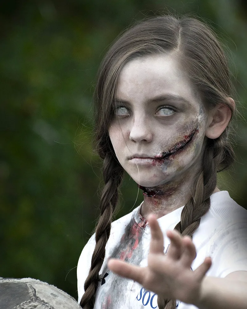 Girl with braids and gray zombie makeup with a stitched scar, reaching toward the camera
